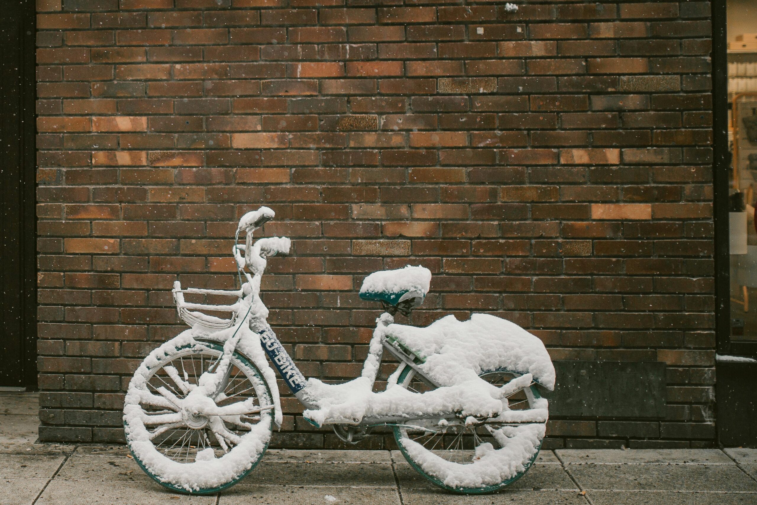 A bicycle covered in snow stands against a brick wall during winter snowfall.